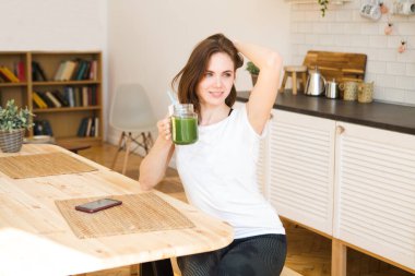 Gülümseyen genç kadın mutfakta yeşil meyve suyu içiyor..
