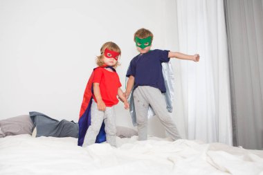 Süper kahraman kostümlü iki anaokulu öğrencisi evdeki yatakta zıplıyorlar.