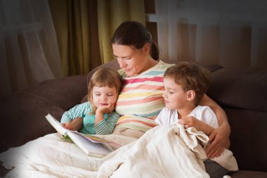 Anne iki çocuğuna kitap okuyor. Kanepede kitap okuyor. Mutlu aile evde birlikte vakit geçirir..