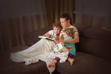 Anne iki çocuğuna kitap okuyor. Kanepede kitap okuyor. Mutlu aile evde birlikte vakit geçirir..