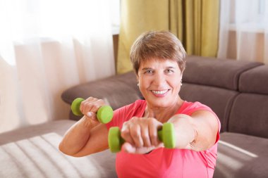 Keyifli yaşlı bayan evde dambıllarla egzersiz yapıyor. Uzaktan antrenman. Evden antrenman yapıyorum. Ev yapımı spor salonu. ev fitness, aktif giyim.