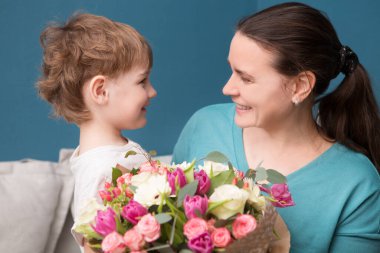 Sevimli küçük çocuk annesine sarılıyor ve onu öpüyor ve mavi arka planda çiçekler sunuyor. Anneler günü, doğum günü.