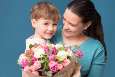 Sevimli küçük çocuk annesine sarılıyor ve onu öpüyor ve mavi arka planda çiçekler sunuyor. Anneler günü, doğum günü.