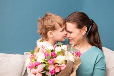 Sevimli küçük çocuk annesine sarılıyor ve onu öpüyor ve mavi arka planda çiçekler sunuyor. Anneler günü, doğum günü.