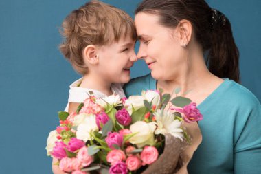 Sevimli küçük çocuk annesine sarılıyor ve onu öpüyor ve mavi arka planda çiçekler sunuyor. Anneler günü, doğum günü.