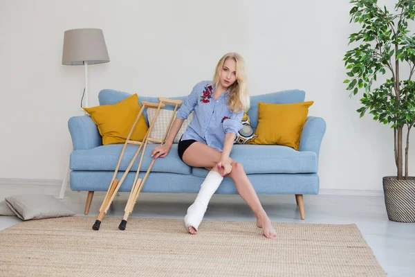 sad Young woman with crutch and broken leg in cast sitting on sofa at home