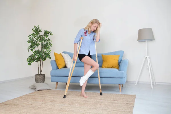 Young woman with crutch and broken leg in cast in the room