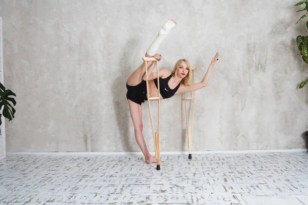 Young woman gymnast in sportswear with crutch and broken leg dances or does gymnastics against the background of a wall