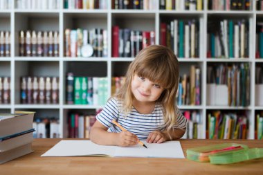 Çocuk evde ders alıyor. Çocuk evde ders çalışıyor. Karantinaya alındı. Corona Virüsü. Liseli kız evde online ders çalışıyor. Evde eğitim, uzak öğrenim. Okul çocuğu arka planda bir kitaplık ile masada çizim yapıyor.