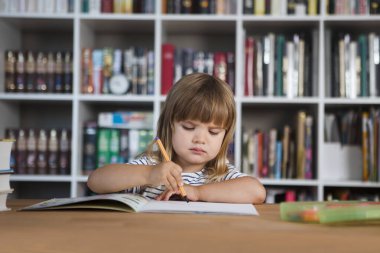 Çocuk evde ders alıyor. Çocuk evde ders çalışıyor. Karantinaya alındı. Corona Virüsü. Liseli kız evde online ders çalışıyor. Evde eğitim, uzak öğrenim. Okul çocuğu arka planda bir kitaplık ile masada çizim yapıyor.