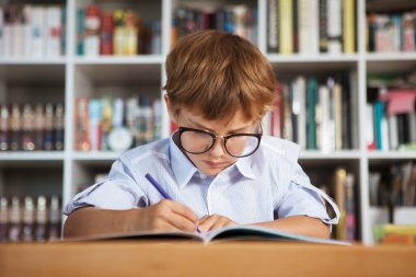 Okula dönüyorum. Okul çocuğu arka planda bir kitaplıkla masada resim çiziyor. 