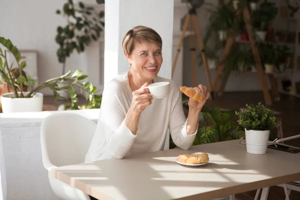 Retired woman eating Croissant and drinking coffee and smiling