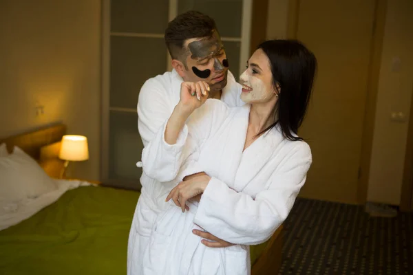 Couple In in white dressing gowns with eye patches relaxing with  clay facial mask. Home spa.