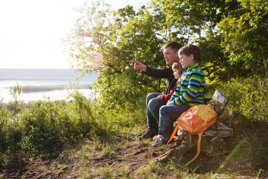 Baba ve sırt çantalı iki çocuk nehir kenarında. Sosyal Mesafe. Dijital detoks. Konaklamalar, hiper-yerel seyahat, aile gezisi, kaçış, doğal çevre