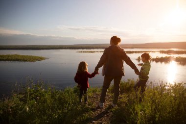 Anne ve iki çocuk nehrin yanında yürüyorlar. Sosyal Mesafe. Karantina sona erdi. Konaklamalar, hiper-yerel seyahat, aile gezisi, kaçış, doğal çevre