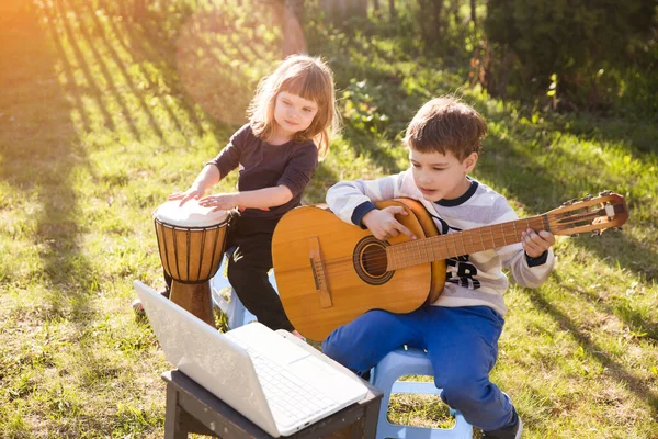 快乐的弟弟和妹妹在户外一起玩乐器 小男孩一边练习吉他 一边在笔记本电脑上看在线课程 — 图库照片