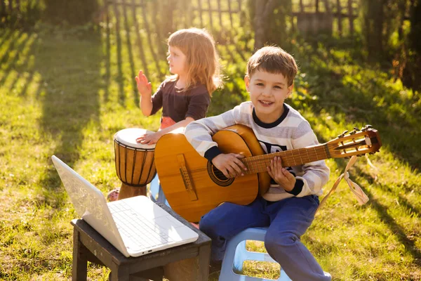 Mutlu çocuklar, erkek ve kız kardeşler dışarıda müzik aletleriyle eğleniyorlar. Gitar çalan ve pratik yaparken online dersler izleyen çocuk. Karantina.