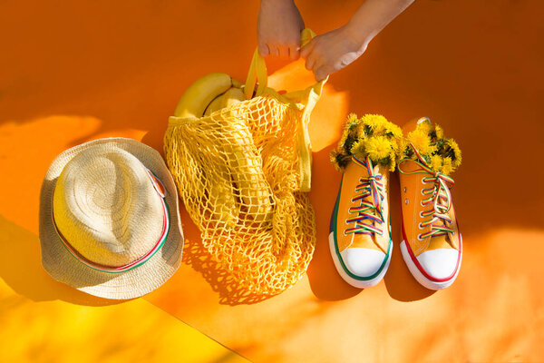 human hand holds mesh shopping bag with bananas and Yellow sneakers flowers dandelions on yellow background with shadows of trees. End of quarantine. Hello summer. summertime