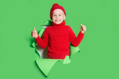 Kırmızı kazaklı ve örgü ören kırmızı şapkalı neşeli çocuk başparmağını kaldırdı ve yırtık kağıt delik duvarında poz verdi. Yırtık kağıdın etkisi. Mutlu Noeller.