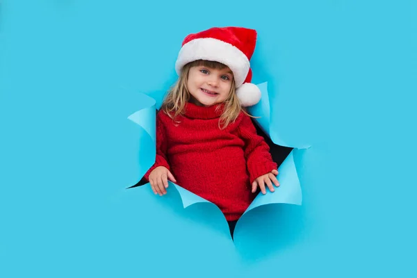 Happy smiling kid in Santa red hat poses through torn paper hole. Effect of torn paper. Christmas sales.