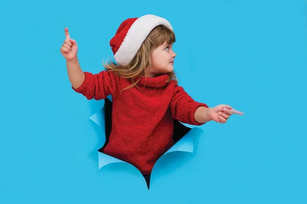 Happy smiling kid in Santa red hat poses through torn paper hole. Effect of torn paper. Christmas sales.