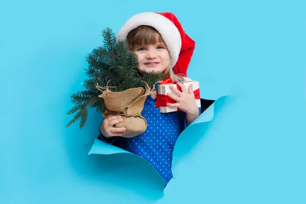 Happy smiling kid in Santa red hat with Christmas tree and gift poses through torn paper hole. Effect of torn paper. Christmas sales.
