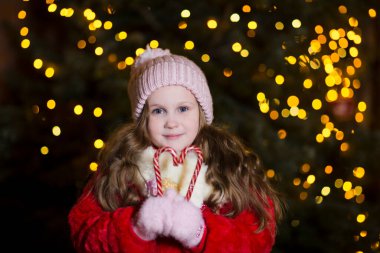 Kış kıyafetleri içinde mutlu bir kız çocuğu. Arka planda ışıklar ve Noel arifesinde açık havada bir Noel ağacı..