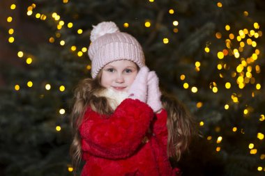 Kış kıyafetleri içinde mutlu bir kız çocuğu. Arka planda ışıklar ve Noel arifesinde açık havada bir Noel ağacı..