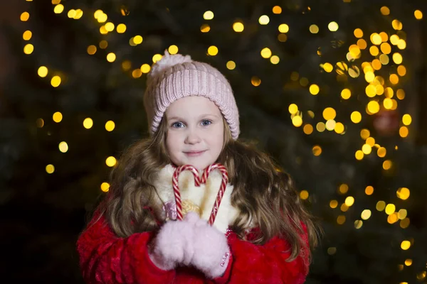 Kış kıyafetleri içinde mutlu bir kız çocuğu. Arka planda ışıklar ve Noel arifesinde açık havada bir Noel ağacı..