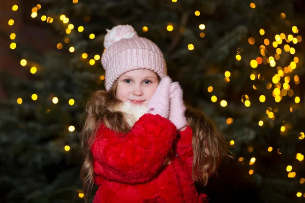 Kış kıyafetleri içinde mutlu bir kız çocuğu. Arka planda ışıklar ve Noel arifesinde açık havada bir Noel ağacı..