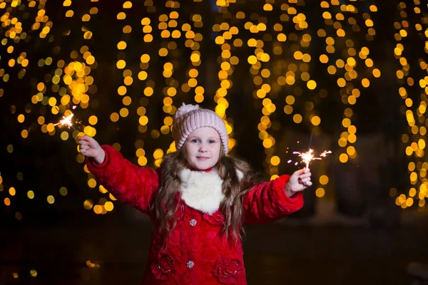 Kış kıyafetleri içinde mutlu bir kız çocuğu. Arka planda ışıklar ve Noel arifesinde açık havada bir Noel ağacı..