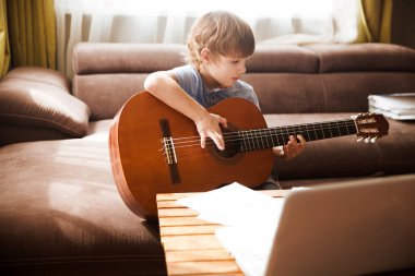 Gitar çalan ve evde pratik yaparken bilgisayardan ders izleyen çocuk. Evde kal. Karantinaya alındı. Çevrimiçi eğitim, online dersler.