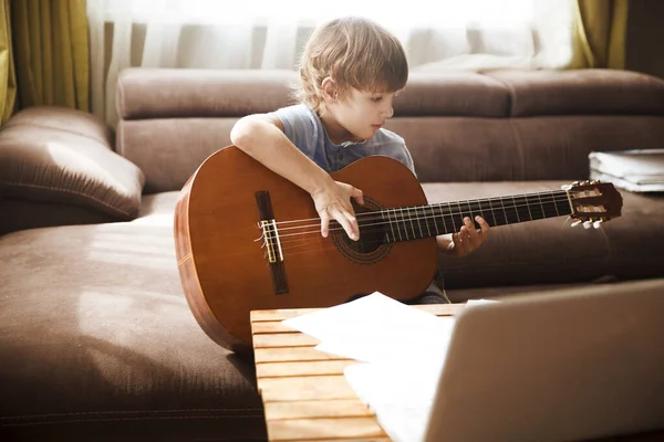Kid boy playing guitar and watching online lessons on laptop while practicing at home. Stay home. quarantine. Online training, online classes.