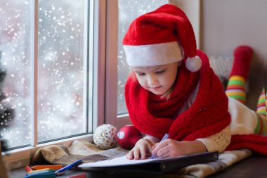 Pencerenin kenarında oturan şirin küçük kız Noel Baba 'ya mektup listesi yazıyor..