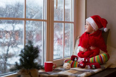 Pencerenin kenarında oturan ve karı seyreden tatlı küçük kız. Noel 'de dekore edilmiş oda. 