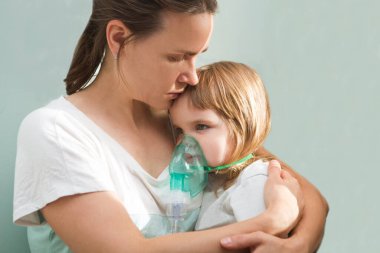 Küçük kız spreyden nebulizörle nefes alıyor. Nefes alma maskesi takmış solunum hastalığına yakalanmış bir çocuk annesinin elinde oturur. Astım hastası.
