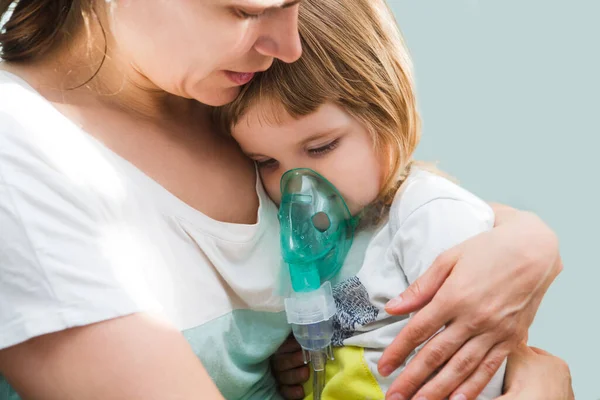 Little child girl inhaling from the inhaler with nebulizer at home. A child  with respiratory disease  in a mask for inhalations sits on hands at the mother. Asthma patien