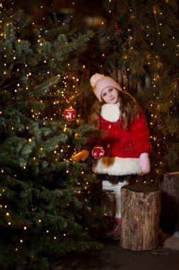 Kış kıyafetleri içinde mutlu bir kız çocuğu. Arka planda Noel süsü, dışarıda Noel arifesinde ise Noel ağacı..
