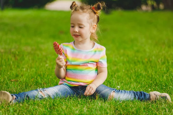 little girl sits on a twine. on the summer grass in the park with a lollipop in hand