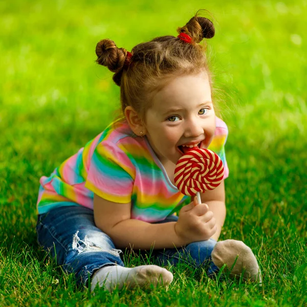 girl with red lollipop sits on the summer grass in the park.