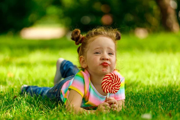 A little girl with a red lollipop lies on the summer grass in the park.