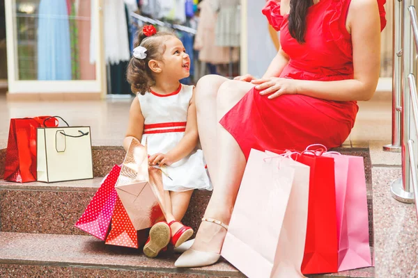 mom with  daughter in dresses are sitting on the steps in the mall with colored bags