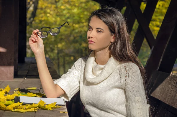 メガネ手で保持して 暖かい秋の午後黄色い葉が付いている公園のベンチに座っている女の子 — ストック写真