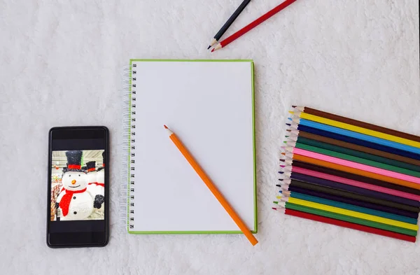notebook, colored pencils for drawing and a phone with a picture of a snowman on a light fluffy background