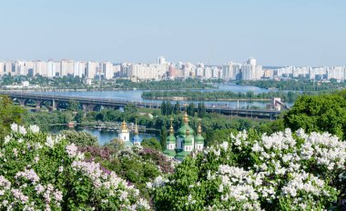 Orthodox church on the background of the river and the city, in the garden of a blossoming lilac, springtime, Ukraine, Kiev clipart