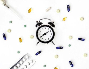 Alarm saati, kapsüllerdeki ilaçlar, arka plandaki şırınga ve haplar.