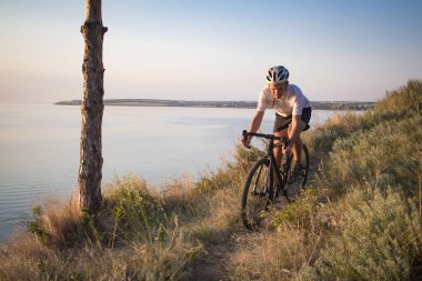 Bisiklet yarışçısı yolculuk yalnız rocky road, göl ve orman arka plan güzel gün batımı sırasında