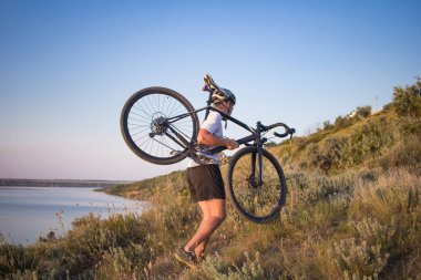 Bisiklet yarışçısı yolculuk yalnız rocky road, göl ve orman arka plan güzel gün batımı sırasında