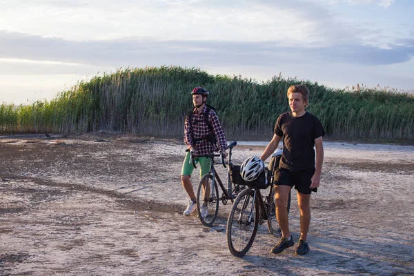 İki genç erkek sırt çantaları ve kask ile bir turne bisikletle binmek açık havada köy yolunda bir bisiklet gezisi
