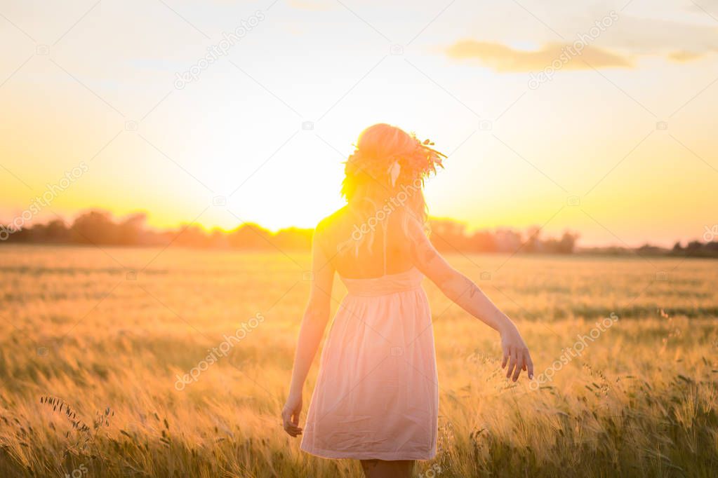 beautiful blonde lady in head flower wreath during sunrise in summer wheat fields